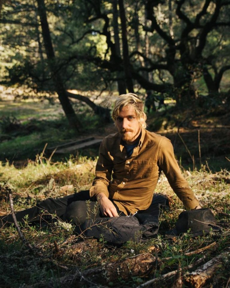 Jay, with bleached hair and a tan button-up shirt, sits in a California forest.