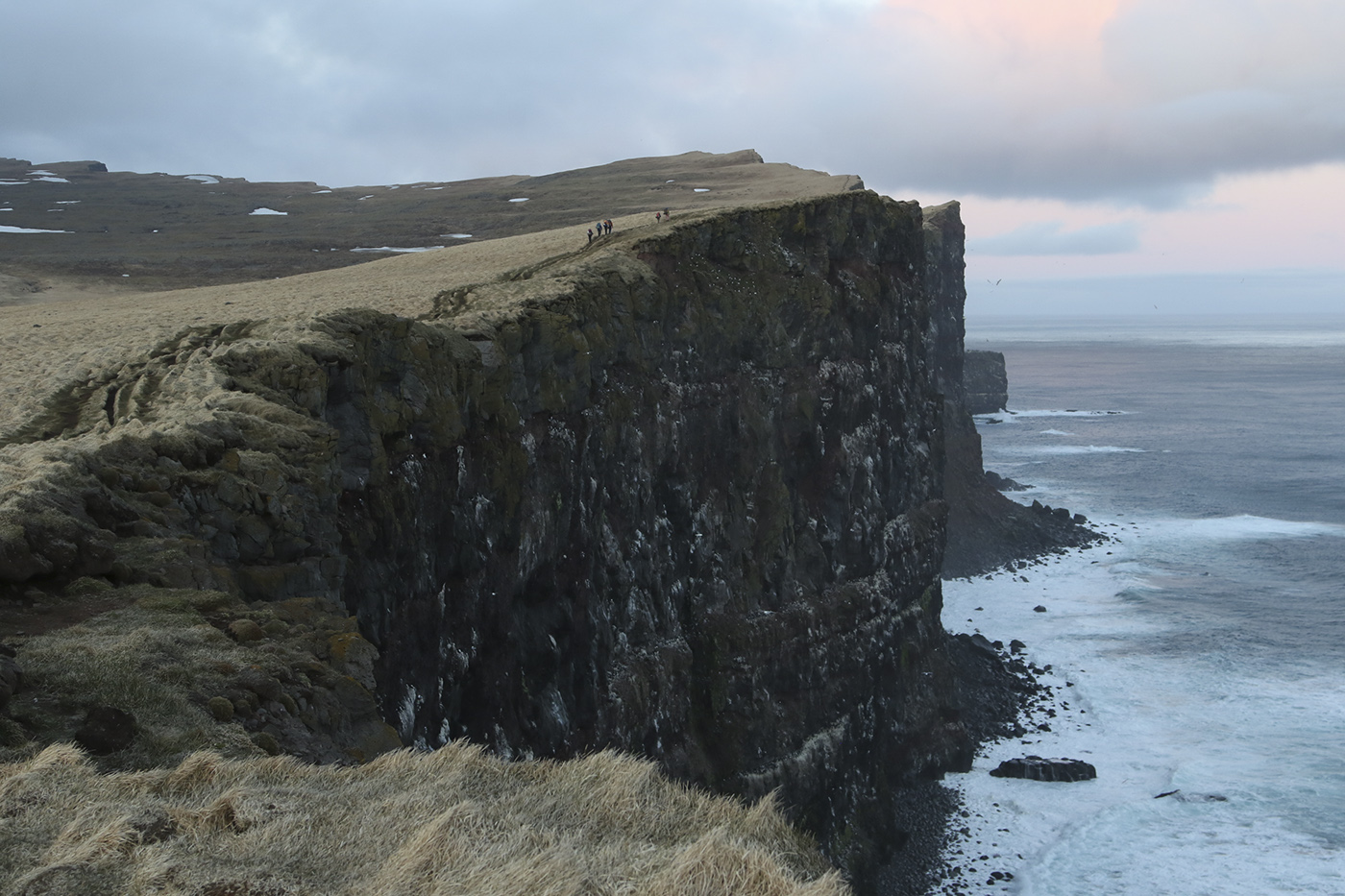 A small group of hikers walk on the edge of tall sea cliffs at arctic midnight.