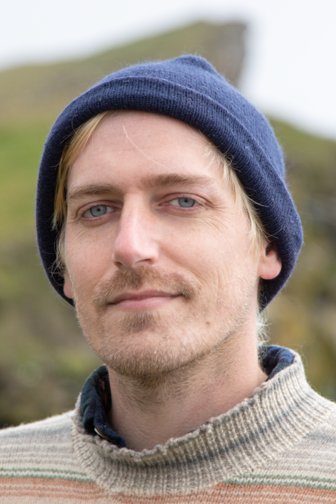 Jay, wearing a blue knit hat and a multicolor sweater, looks directly at the viewer. He is white, has light blue eyes and bleach blonde hair pokes out in places from his hat.